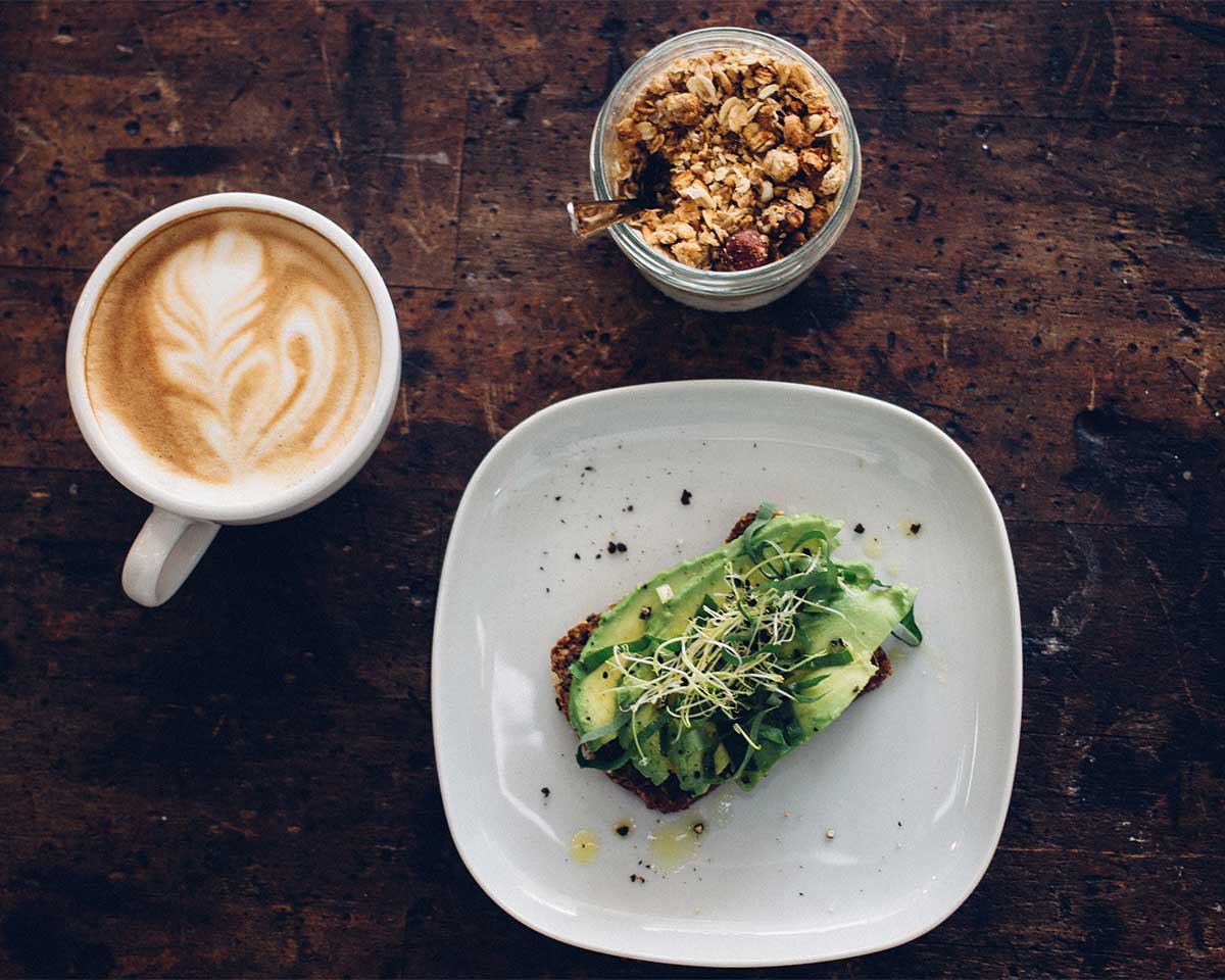 an appetizing shot of a plant based meal - avocado toast with sprouts and cracked pepper, a latte, and a granola parfait