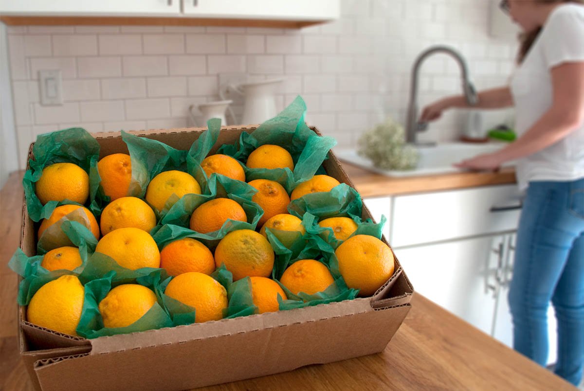 SweeterSorts citrus packaged in box sitting on kitchen counter with girl in background washing hands at the sink