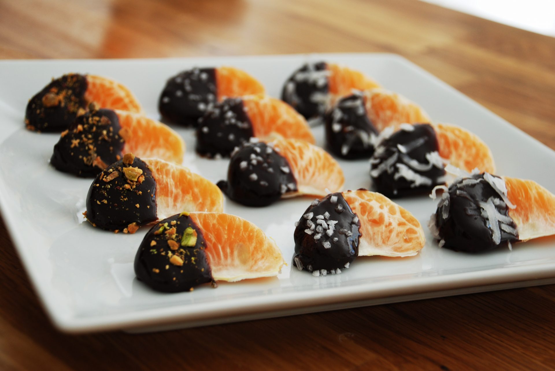 Chocolate dipped tangerines sprinkled with nuts on white plate on wooden countertop